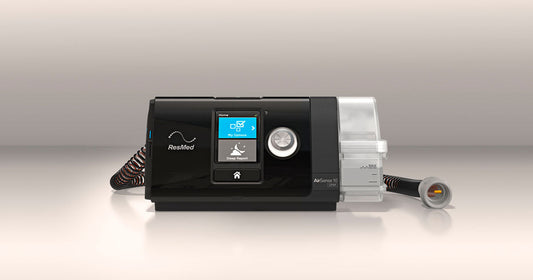 Black AirSense 10 AutoSet CPAP machine on a white counter against a white wall. Machine screen is lit up to display the Home menu of the AirSense 10 machine.